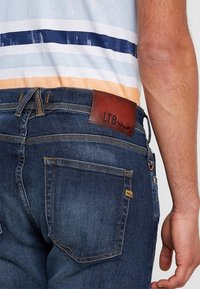 Džínové kalhoty s tmavě modrým praním, s hnědou koženou nášivkou s nápisem "LTB." Viditelné zadní kapsy a standardní švy.