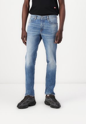 Homme portant un jean bleu clair délavé, des baskets noires et un t-shirt noir, debout devant un fond blanc.
