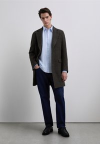 Abrigo marrón de lana con diseño entallado, camisa azul claro abotonada debajo, pantalones vaqueros oscuros y zapatos negros. Fondo liso.