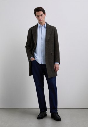 Abrigo marrón de lana con diseño entallado, camisa azul claro abotonada debajo, pantalones vaqueros oscuros y zapatos negros. Fondo liso.