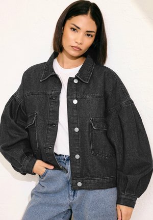 Frau mit glatten dunklen Haaren trägt eine übergroße schwarze Jeansjacke über einem weißen Hemd und blauen Jeans, posiert vor einer schlichten hellen Wand.