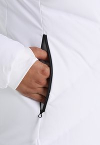 Eine Hand teilweise in der unzipped Tasche mit schwarzem Rand einer weißen Jacke, wobei ein weißer Bund sichtbar ist.
