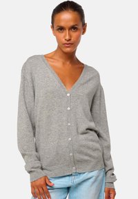 Femme portant un cardigan gris clair à boutons avec un décolleté en V profond et un jean bleu clair, se tenant contre un fond uni clair.