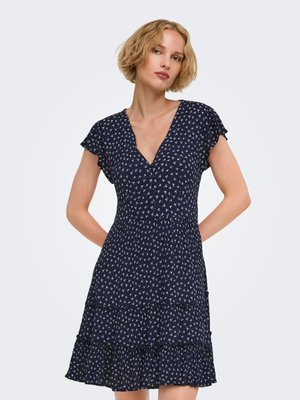 Femme blonde aux cheveux courts portant une robe bleu marine à imprimé floral, manches courtes et col en V, debout avec les mains dans le dos.