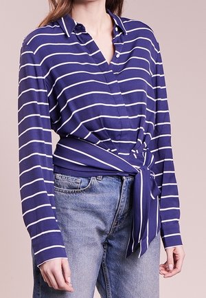 Frau trägt ein blau-weiß gestreiftes Wickelshirt mit langen Ärmeln, kombiniert mit hellblauen Jeans.