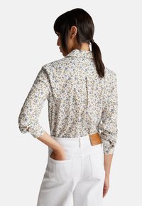 Donna con capelli scuri raccolti in una coda di cavallo, che indossa una camicia bianca a maniche lunghe con motivo floreale infilata in pantaloni bianchi a vita alta, con una mano nella tasca posteriore.