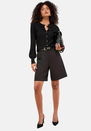 Schwarzes geripptes Langarmshirt mit Knöpfen, hochgeschnittene schwarze Shorts und schwarze High Heels. Eine einfache schwarze Handtasche vervollständigt das Outfit.