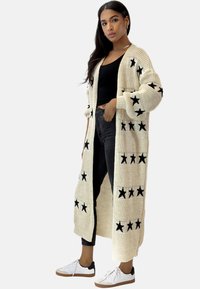 Langer, cremefarbener Cardigan mit schwarzen Sternmustern, gestrickt, mit übergroßen Ärmeln und offenem Vorderteil. Kombiniert mit schwarzen Hosen und Turnschuhen.