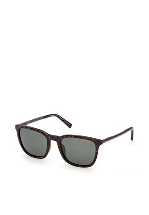 Quadratisch geformte Sonnenbrille mit dunkelgrünen Gläsern und braunem Schildpattgestell, die auf einer weißen Oberfläche liegt.