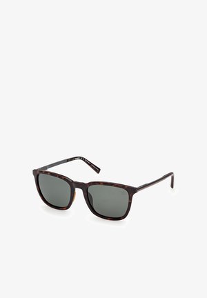 Quadratisch geformte Sonnenbrille mit dunkelgrünen Gläsern und braunem Schildpattgestell, die auf einer weißen Oberfläche liegt.