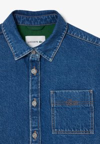 Denim shirt in medium blue featuring a button-down collar, chest pocket with embroidered logo, and contrasting green inner lining.