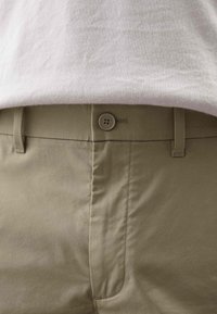 Pantalones caquis con frente plano, cierre de botón y trabillas para cinturón. Textura de tela suave con detalles de costura mínimos.