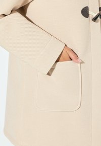 Béžový texturovaný kabát s nášivkou na boku a zapínáním na toggle. Materiál vypadá tkaný a odolný.