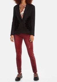 Mujer con blazer negro a medida con paneles de encaje, blusa estampada, pantalones ajustados rojos con adornos y zapatillas negras con cordones estampados.