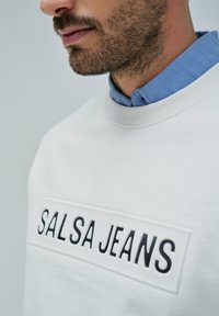 Sudadera blanca con un logo texturizado de "SALSA JEANS" en negro. Llevada sobre una camisa azul clara, con un cuello redondo y puños acanalados.