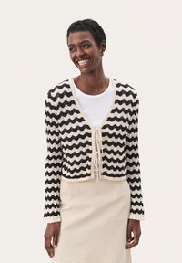 Cardigan tricoté avec un motif ondulé noir et crème, col en V, fermeture à nouer et bords festonnés. Porté sur une chemise blanche et une jupe beige.