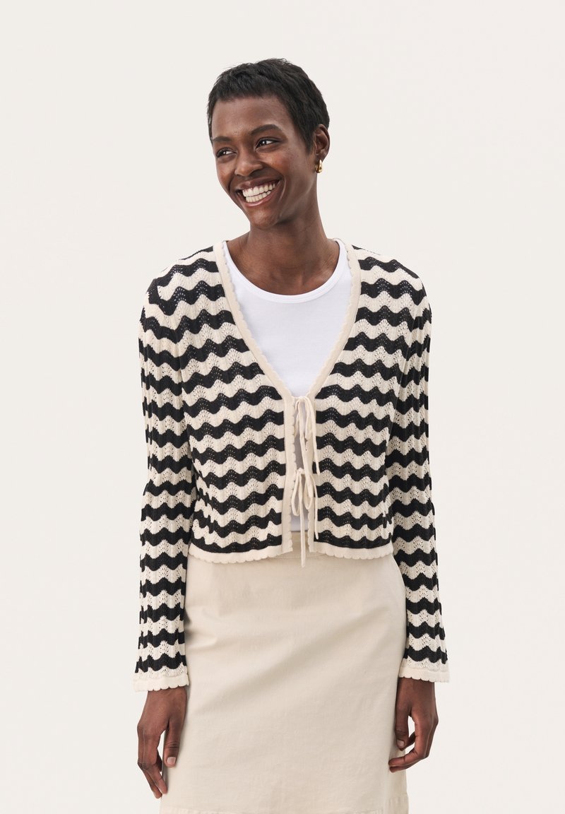 Cardigan tricoté avec un motif ondulé noir et crème, col en V, fermeture à nouer et bords festonnés. Porté sur une chemise blanche et une jupe beige.