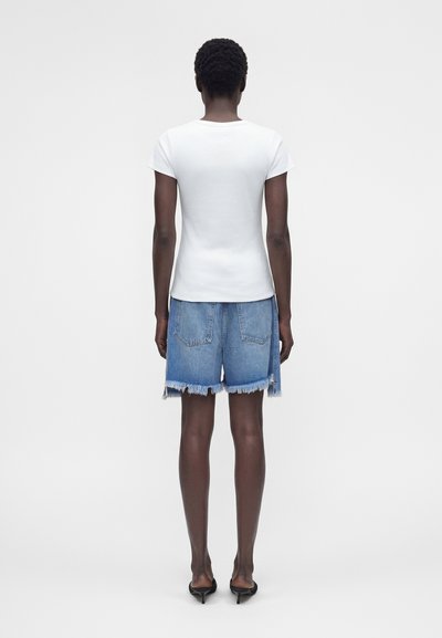 Άτομο που στέκεται με την πλάτη στην κάμερα, φοράει λευκό απλό εφαρμοστό μπλουζάκι, ξεφτισμένες μπλε τζιν κοντές παντελόνες και μαύρα σανδάλια με τακούνι.