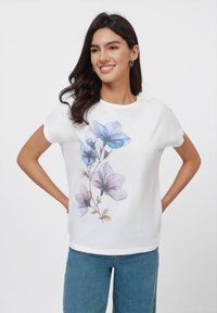 T-shirt en coton blanc avec un motif floral dans des nuances de bleu et de violet, dotée de manches courtes et d'un col rond, associée à un jean bleu.