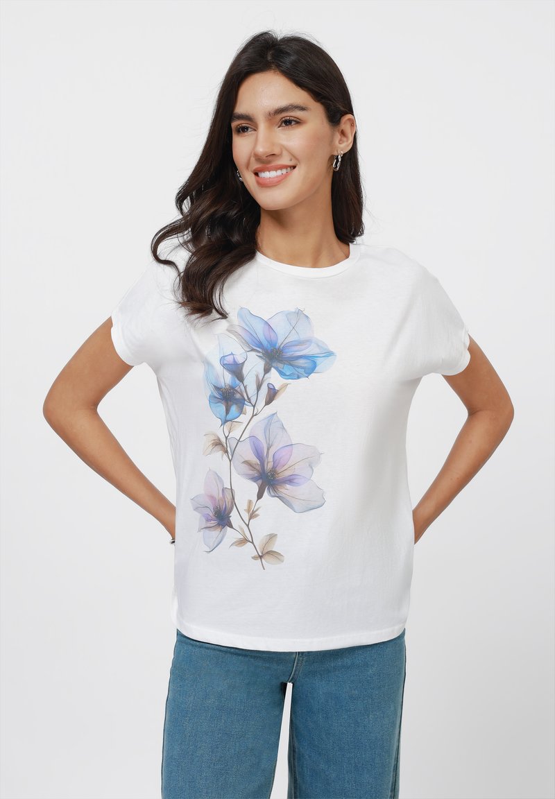 T-shirt en coton blanc avec un motif floral dans des nuances de bleu et de violet, dotée de manches courtes et d'un col rond, associée à un jean bleu.