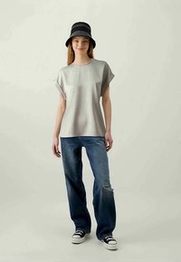 Top cinza de mangas curtas com um corte solto, combinado com jeans azuis desgastados e um chapéu preto de balde. Ténis preto e branco completam o visual.