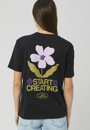 Femme aux longs cheveux bruns portant un t-shirt noir avec une grande fleur violette et les textes "COMMENCEZ À CRÉER" et "ARRÊTEZ DE FAIRE DÉFILER" au dos.