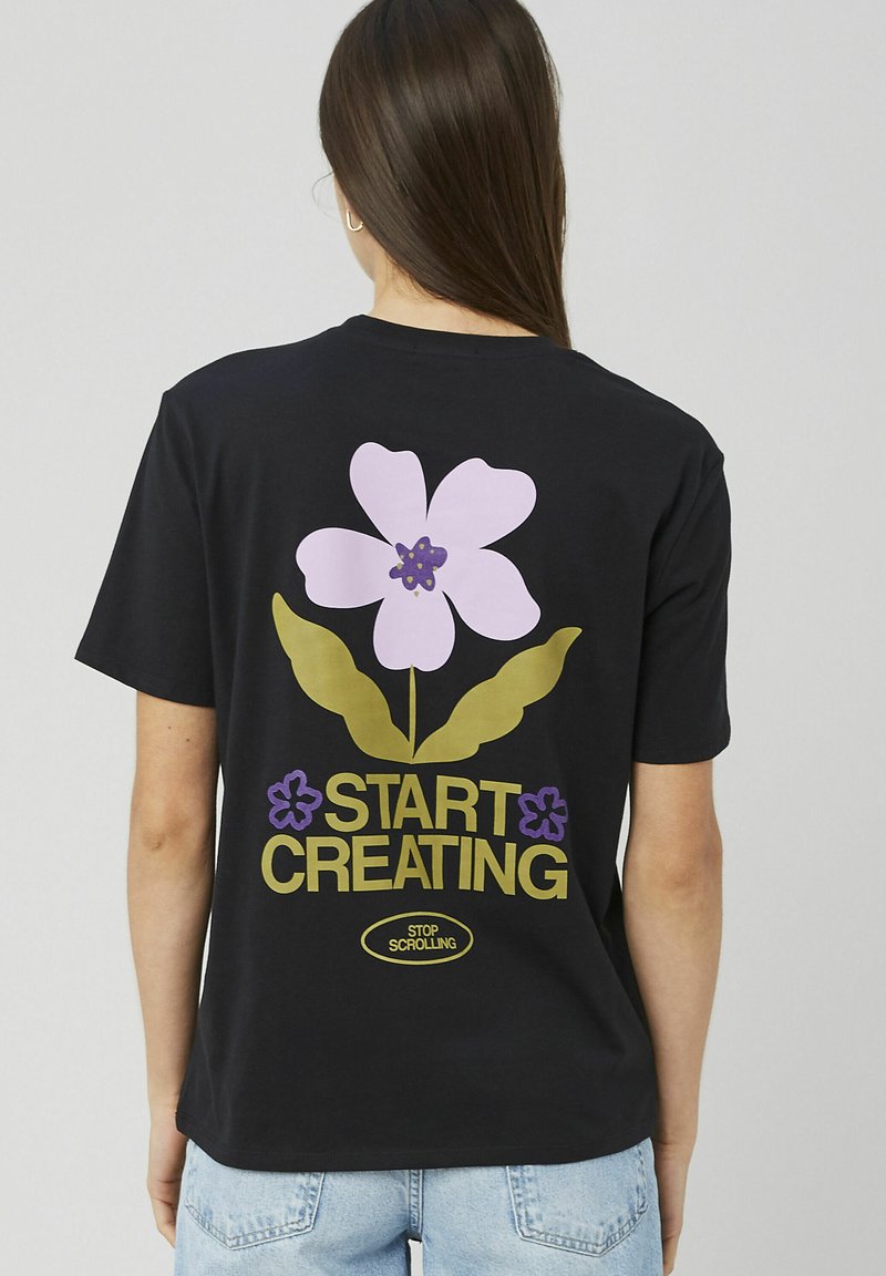 Femme aux longs cheveux bruns portant un t-shirt noir avec une grande fleur violette et les textes "COMMENCEZ À CRÉER" et "ARRÊTEZ DE FAIRE DÉFILER" au dos.