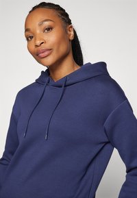 Navy blå hettegenser med snøring i hetten, med en myk tekstur og avslappet passform. Enkel design uten synlige mønstre eller detaljer.