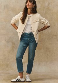 Cremefarbene Blazer mit goldenen Knöpfen, weißem T-Shirt, dunkelblauen Jeans im geraden Schnitt und weißen Sneakern mit schwarzen Streifen.