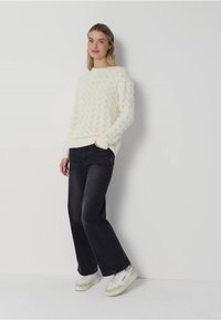 Weißer gestrickter Pullover mit einem strukturierten Muster, kombiniert mit schwarzen Schlagjeans und weißen Sneakers mit beigen Akzenten.