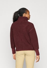 Bordeaux fleece genser med høy krage, teksturert overflate og lange ermer. Kombinert med lyse bukser, som viser en avslappet passform.