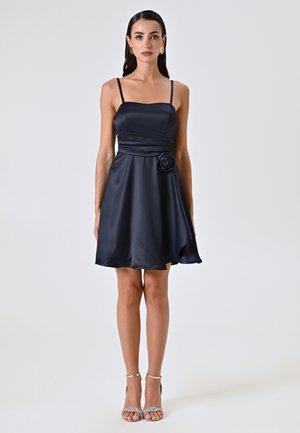 Robe en satin marine avec un corsage ajusté, des bretelles spaghetti et un accent floral �à la taille. Jupe évasée avec un ourlet inégal. Talons argentés.