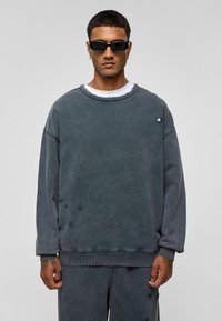 Grijze oversized hoodie van katoen met verouderde details, ronde hals, ribgebreide mouwen en zoom. Draag het over een wit T-shirt.