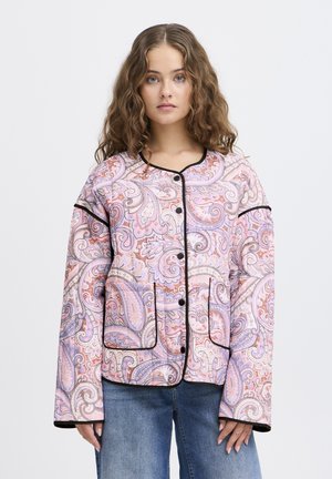 Jeune femme aux cheveux ondulés portant une veste à boutons à motif paisley pastel et un jean bleu sur un fond clair uni.