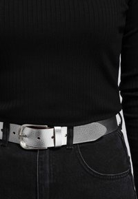 Silverfärgad läderbälte med texturerad yta, med en metallspänne och flera justeringshål, som bärs över en svart ribbad topp och mörka jeans.