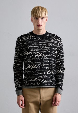 Schwarzer Pullover mit weißem, geschwungenem Schriftmuster. Gerippter runder Halsausschnitt und Bündchen. Das Model trägt ihn mit beigen Hosen. Strukturierter Strickstoff.