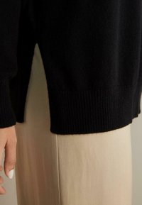 Svart stickad tröja med ribbade muddar och nederkant, med en sidolikning. Kombinerad med beige byxor som har en slät yta.