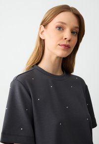 Donkergrijze blouse met korte mouwen en een ronde halslijn, voorzien van verspreide zilveren studs als accenten op de stof. Zachte textuur.