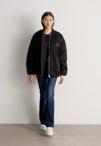 Jauna sieviete pastaigājas iekštelpās, valkājot melnu polsterētu jaku, tumšas platām kājām džinsus un baltus sporta apavus, atrodoties pret vienkāršu balto sienu.