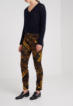 Femme portant un pull noir à col en V pelucheux, un pantalon noir à motifs dorés et des chaussures noires sans lacets, debout devant un fond blanc.