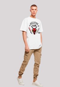 White cotton t-shirt featuring a vibrant red and black graphic with a skull and "HELLFIRE CLUB" text, paired with beige cargo pants and white sneakers.