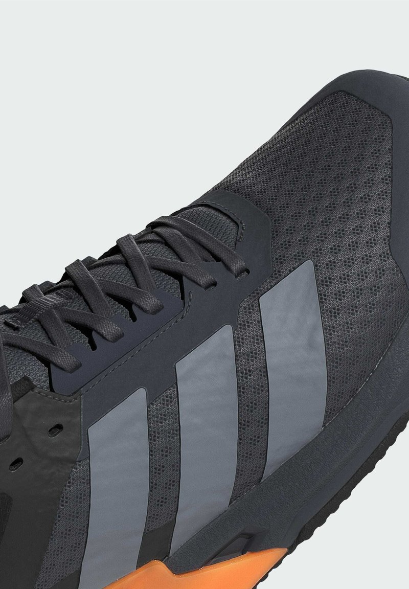 Sportowe buty z ciemnoszarym siateczkowym wierzchem, teksturowanymi akcentami syntetycznymi, trzema odblaskowymi paskami i pomarańczową podeszwą. Sznurówki są ciemnoszare.