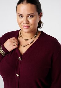 Bordeaux cardigan met een V-hals, voorzien van goudkleurige knopen. Gekleed met gelaagde goudkleurige kettingen en armbanden.