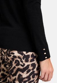 Fekete kötött pulóver bordázott aljjal és három arany árnyalatú gombbal díszített mandzsettával, leopárdmintás leggingszel párosítva.