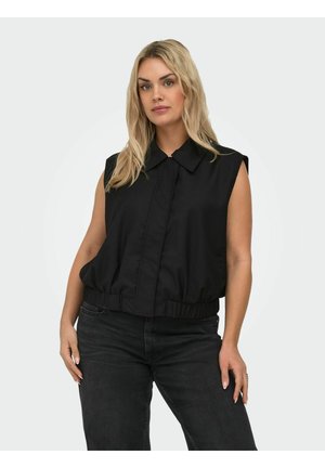 Blond naine kannab varrukateta musta kraega pealset ja musti teksapükse, seisab käed puusadel ühevärvilise tausta ees.