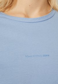 Närbild av en blå t-shirt med rund halsringning med blekt "Marc O'Polo DENIM"-logotyp på bröstet, som bärs av en person med blont hår.