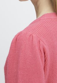 Suéter de punto rosa con un acabado texturizado. Presenta un cuello redondo y detalles fruncidos en el hombro. Material suave y ligero.