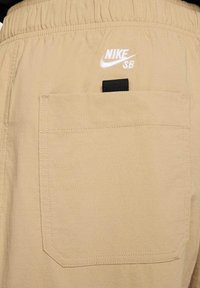 Beżowe szorty z elastycznym paskiem w talii, białym haftowanym logo "Nike SB" oraz prostokątną tylną kieszenią z czarnym akcentem.