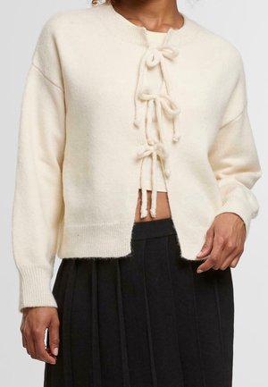 Woman wears a cream cardigan with three front bow tie closures and a black pleated skirt, standing against a plain background.