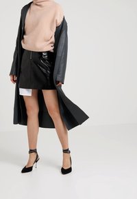 Jupe mini en simili cuir noir avec une fermeture éclair à l'avant, associée à un col roulé moelleux rose clair et à un long manteau en cuir noir. Talons noirs à bride cheville.